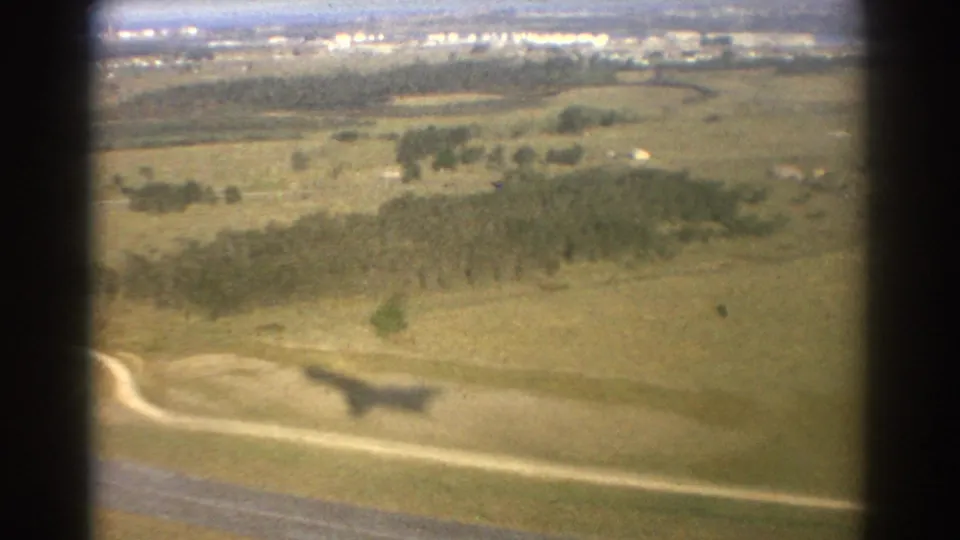 1967 Brisbane aerial view from a moving aircraft, looking over open grassland, tree clusters, road curves, and distant buildings.