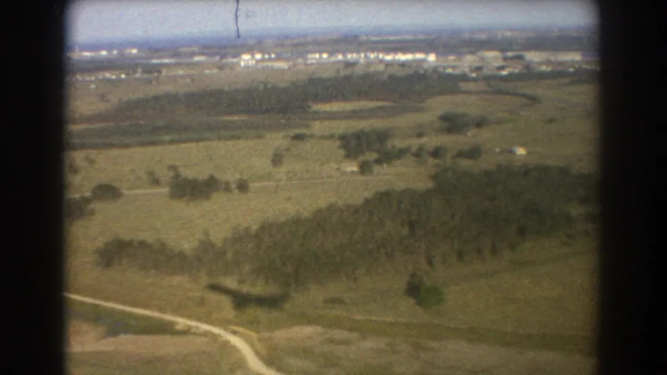 1967 Brisbane home-movie aerial frame showing open fields, roads, tree lines, distant development, and a plane shadow on the ground.