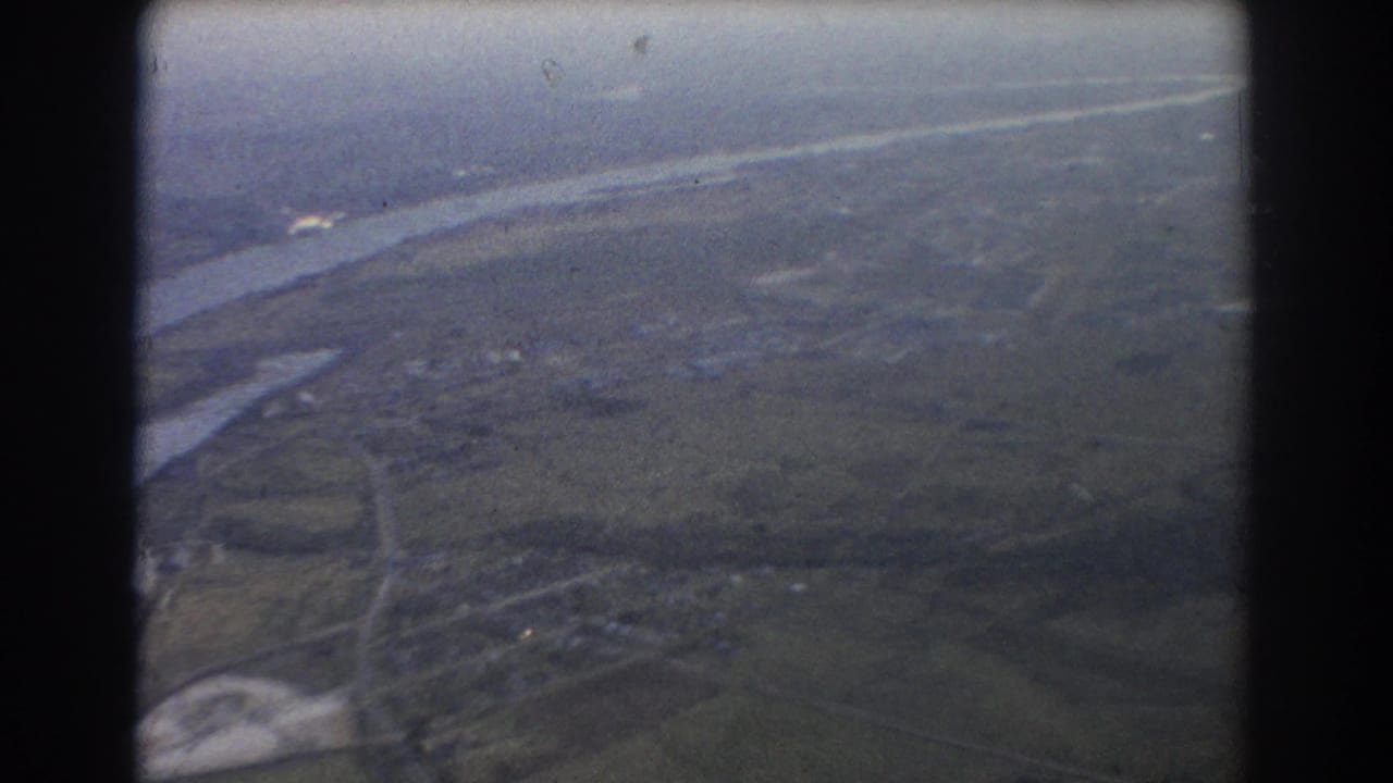 Soft aerial view of river channels, open land, roads, and small building clusters through film grain.