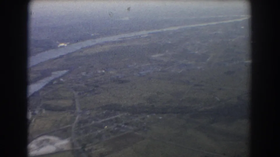1967 Brisbane-area aerial view with a broad river corridor, fields, roads, and distant settlement softened by haze and 8mm grain.