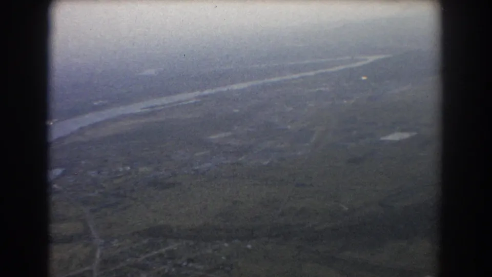 1967 Brisbane-area home-movie aerial frame showing river water, rural fields, road traces, and distant buildings from a passenger viewpoint.