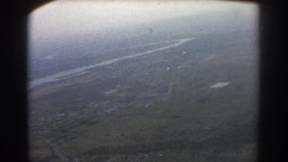 A soft 8mm aerial view from an airplane window, with terrain below and a winding water feature visible through haze.