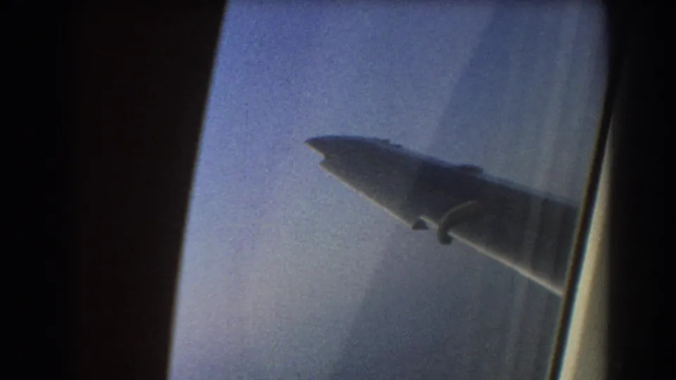 1967 8mm airplane-window view showing the aircraft wing outside against a pale blue sky and window reflections.