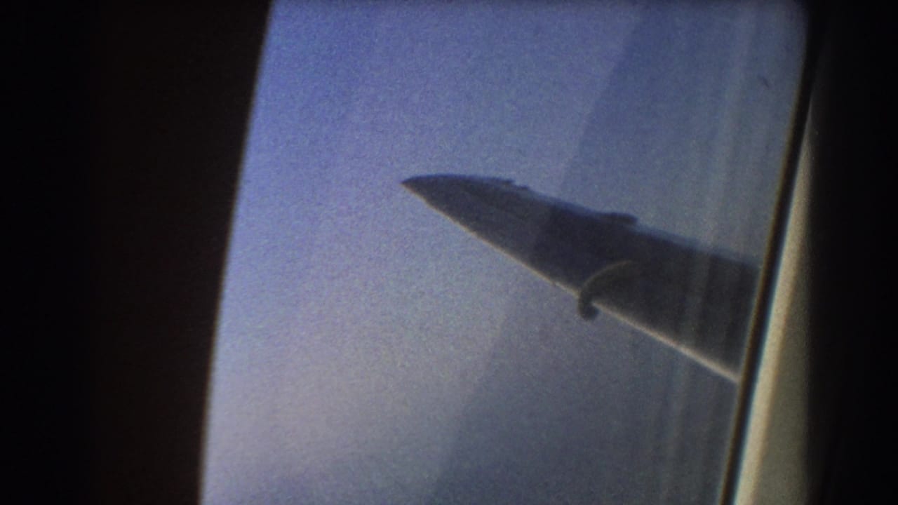 1967 8mm travel footage showing an airplane wing through a passenger window, with sky haze and soft film grain.