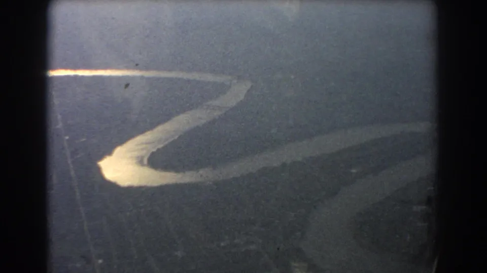 Aerial 8mm view near Brisbane showing a pale river bend against darker terrain, framed by the aircraft window.