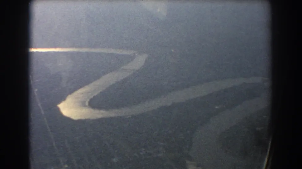 1967 home-movie frame from a plane window, with a glowing river curve and muted rural ground below.