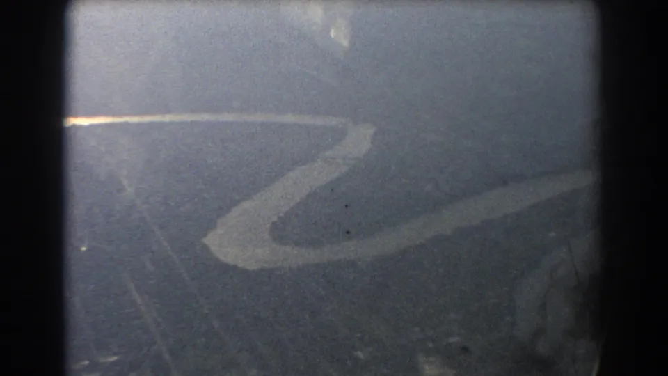 1967 8mm aerial frame with a pale curving waterway or inlet, soft focus, dust, and heavy dark film-gate borders.