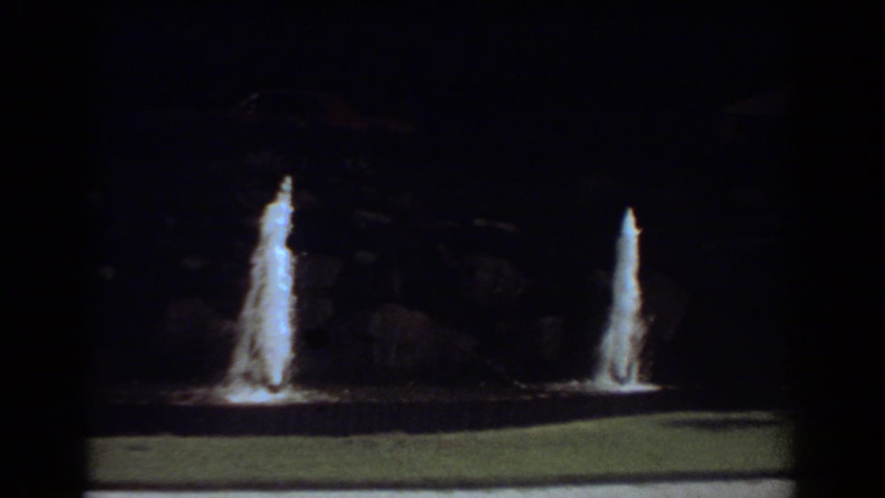 Two lit water jets rise from a low fountain basin in a dark outdoor setting.