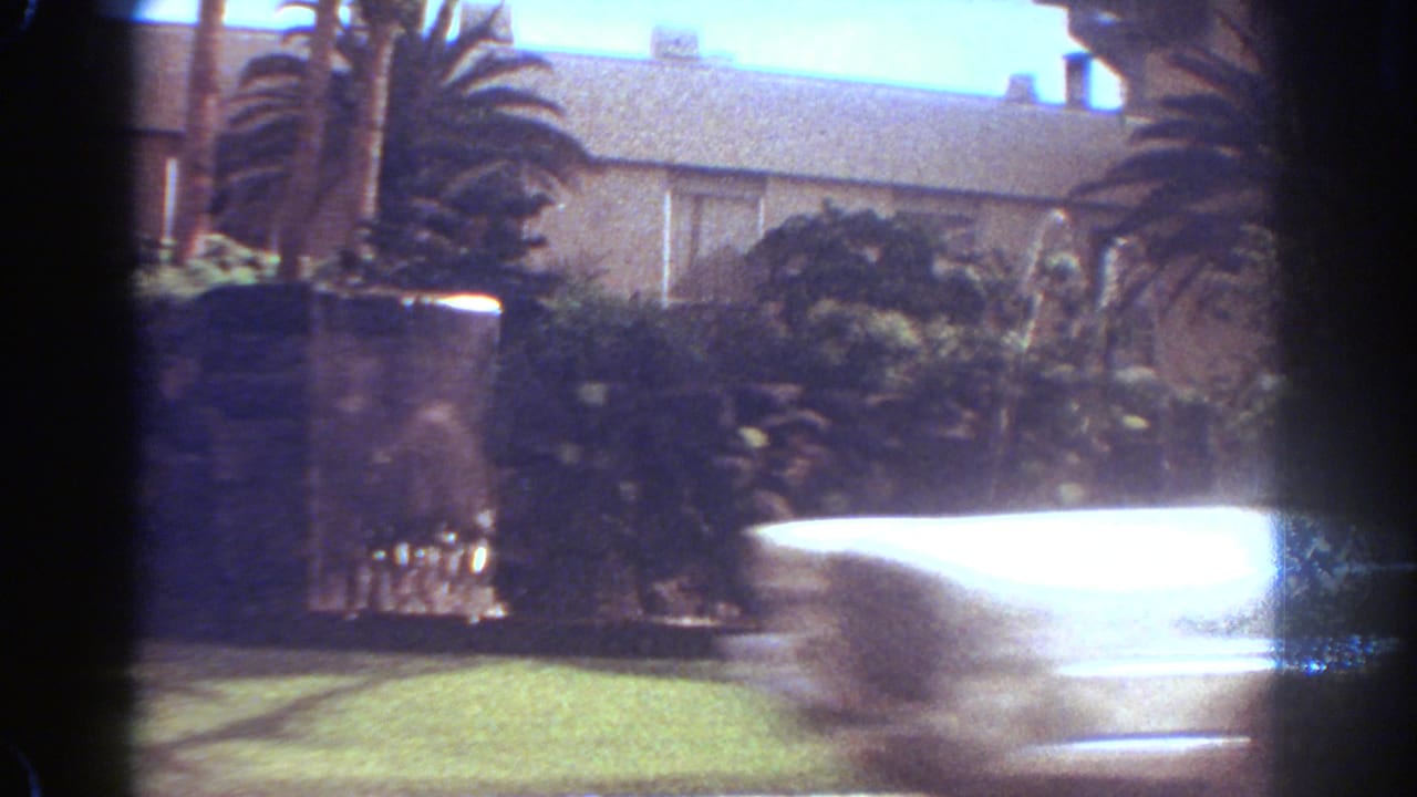 1967 Fort Lauderdale home-movie frame showing the fountain area with bright water spray beside palms, shrubs, and lawn.