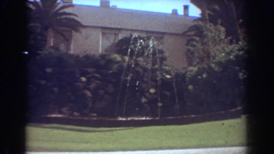 1967 Fort Lauderdale frame of a residential exterior with palms, dense landscaping, lawn, and a visible front-yard fountain.