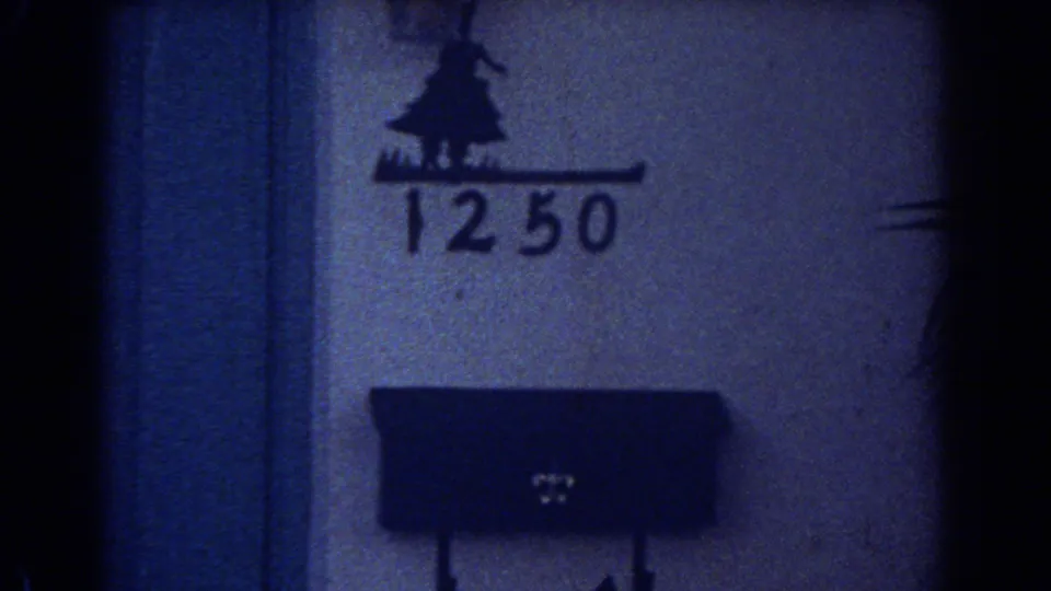 Close 1967 home-movie frame showing the number 1250 on a wall above a dark mailbox, with heavy grain and shadow.
