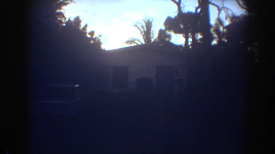 1967 Fort Lauderdale home-movie frame showing a white house, lawn, trees, and a parked car in strong evening backlight.