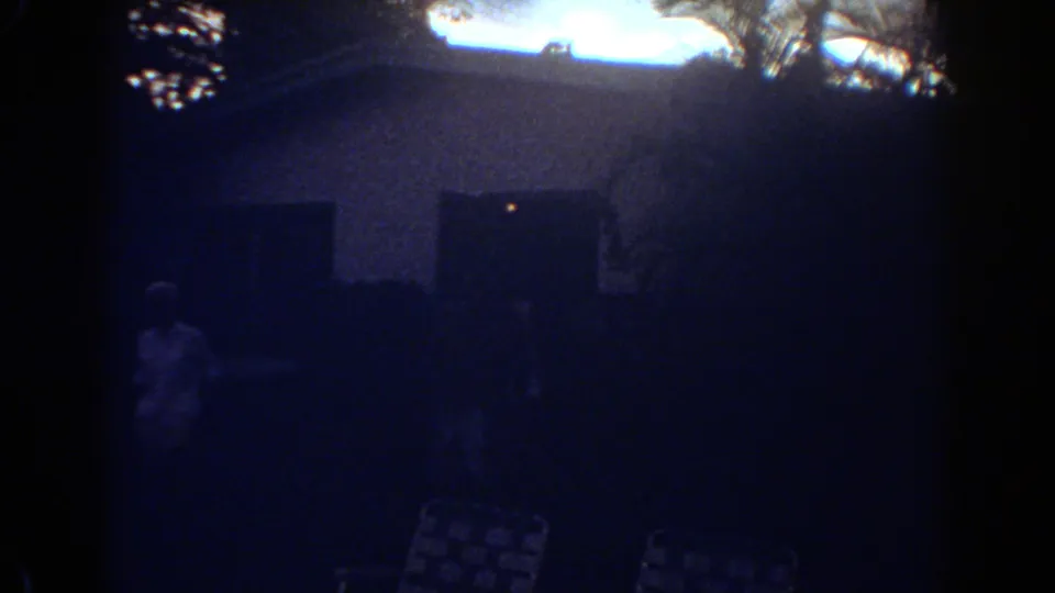 1967 Fort Lauderdale home-movie frame showing a dark house entrance, a person in silhouette, lawn chairs, and palm shapes against dusk light.