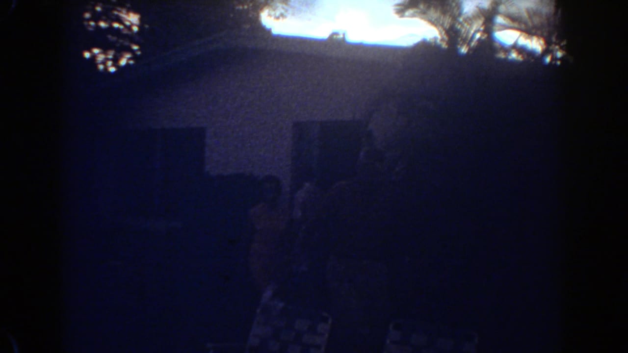 1967 Fort Lauderdale frame with a silhouetted woman in a patterned dress near the house entrance, seen through grainy dusk exposure.