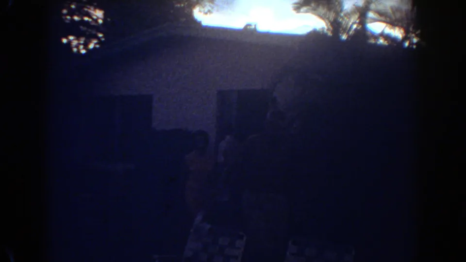 1967 Fort Lauderdale frame with a silhouetted woman in a patterned dress near the house entrance, seen through grainy dusk exposure.