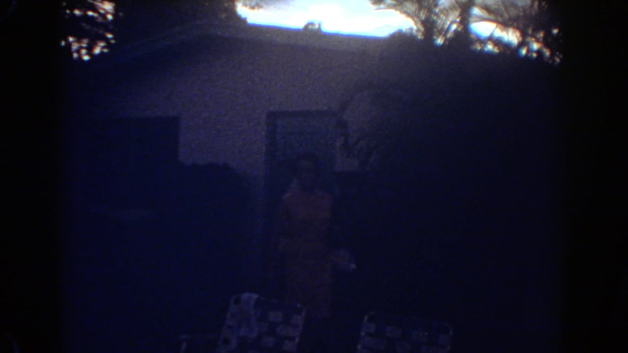 Grainy 8mm frame from Fort Lauderdale in 1967 showing a dim house exterior, a person near the entrance, and patterned lawn chairs.