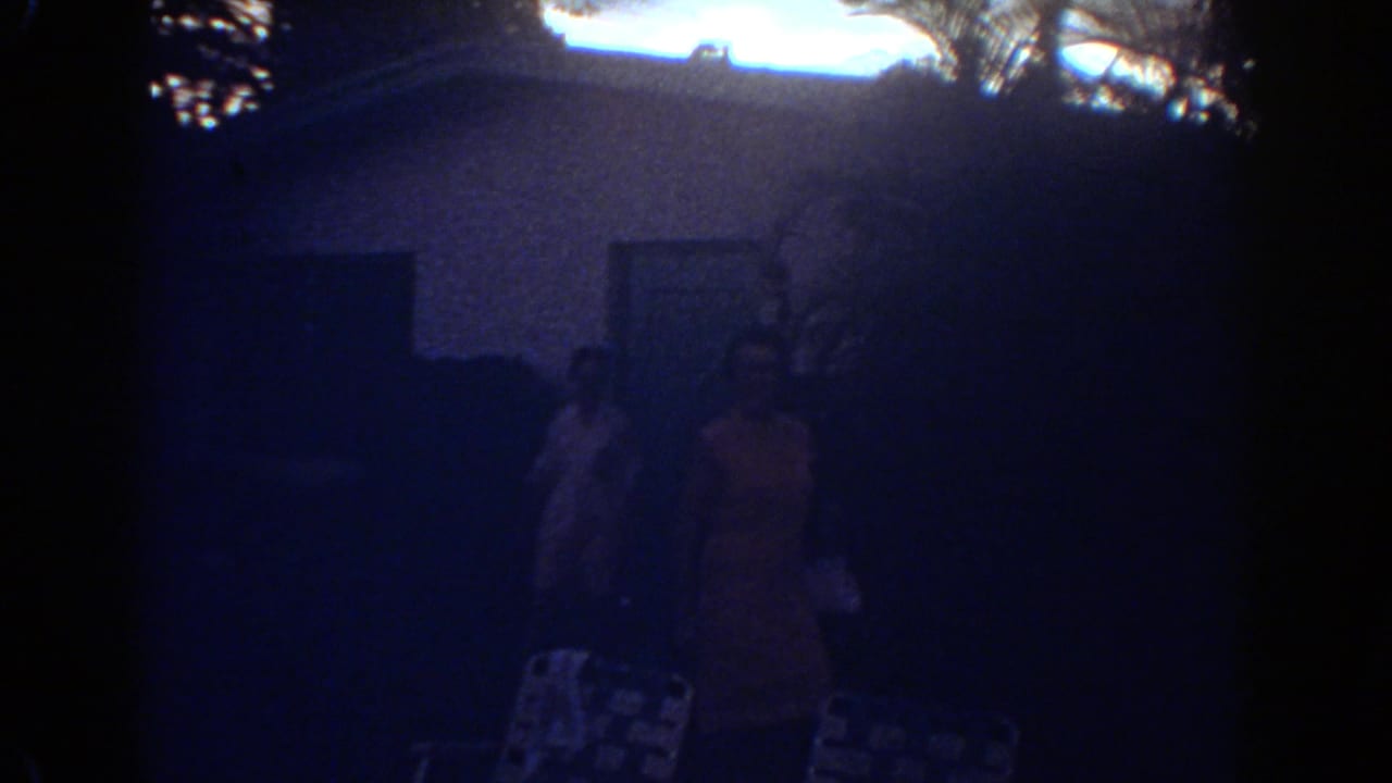 Very dark frame with faint figures near a building in strong shadow.