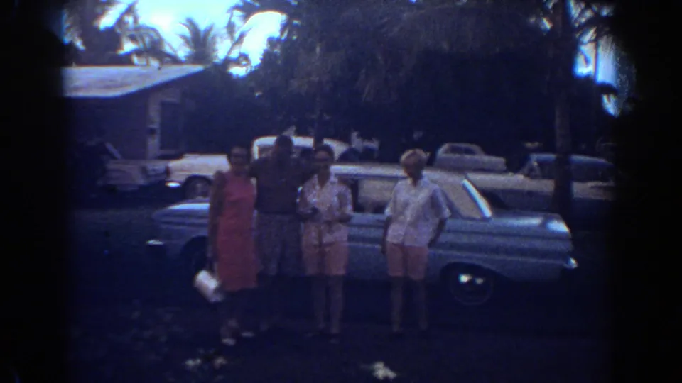 Four adults in 1967 Fort Lauderdale pose near a light blue car with palm trees and a house behind them.