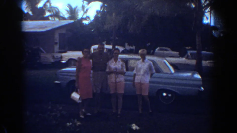 Four adults face the camera near a light blue car in Fort Lauderdale, with palms and parked cars behind them.