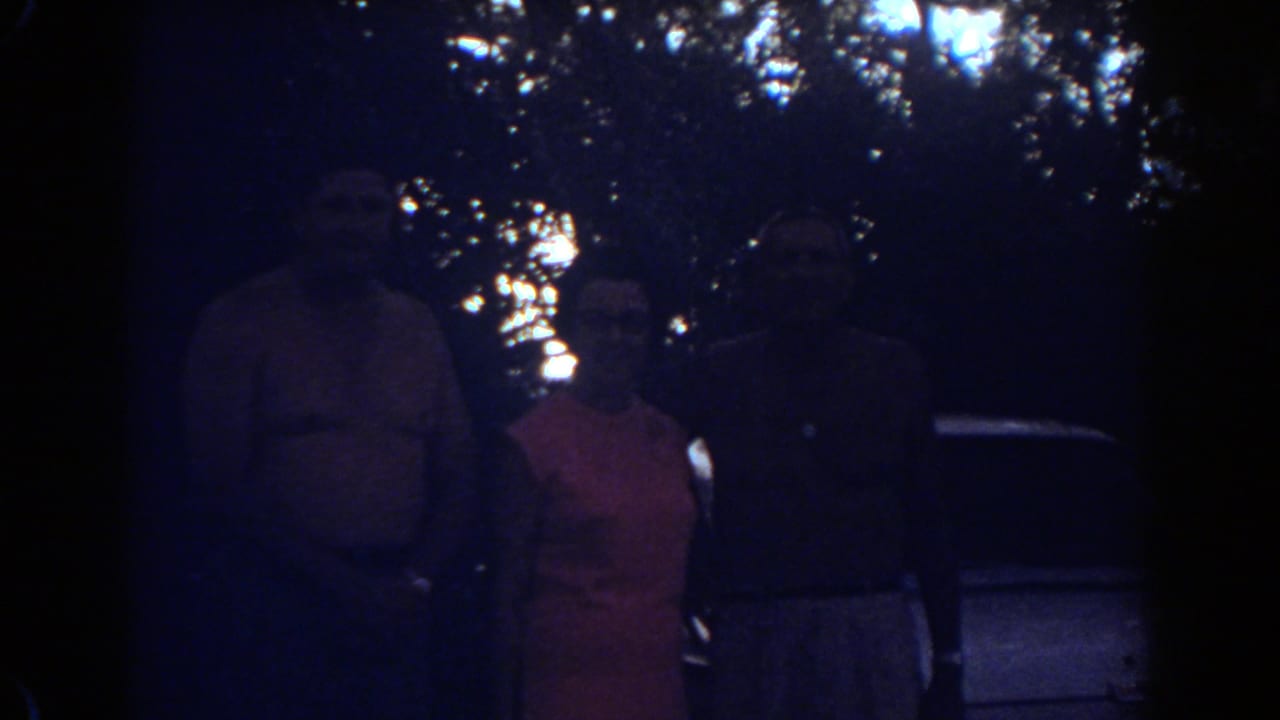 Three adults pose in a dim 1967 Fort Lauderdale home-movie frame, with foliage and a car edge visible behind them.