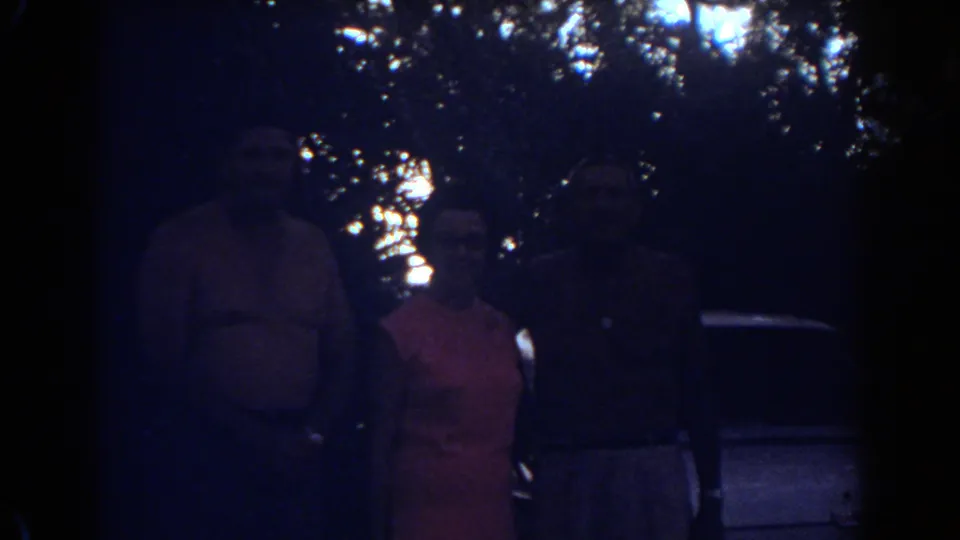 Three adults pose in a dim 1967 Fort Lauderdale home-movie frame, with foliage and a car edge visible behind them.