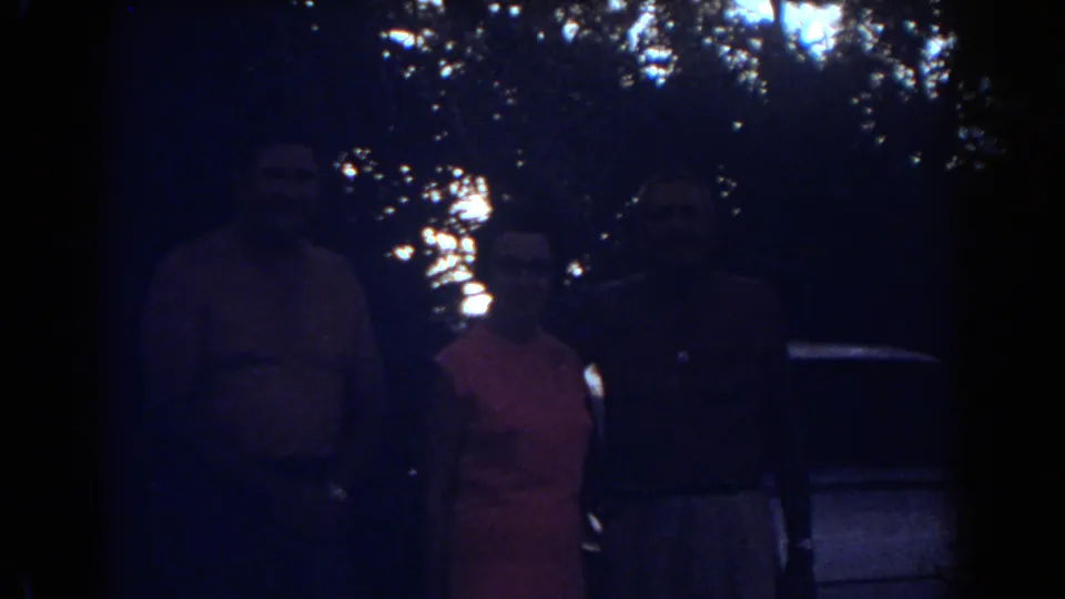 Dark 8mm frame of three adults standing under trees, with faces and clothing partly obscured by shadow in a Fort Lauderdale home-movie scene.