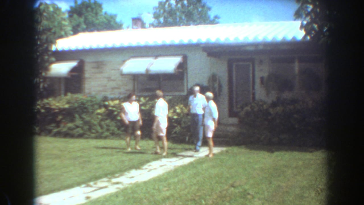 Four people stand on a front lawn near a stone path outside a single-story house.