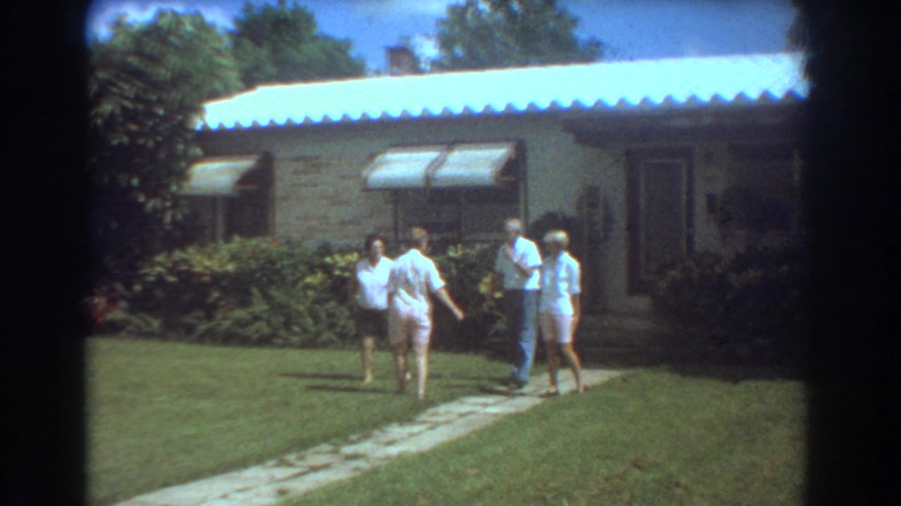 In a 1967 Fort Lauderdale yard, four people walk and turn along a stone path before a single-story house with shrubs and awnings.