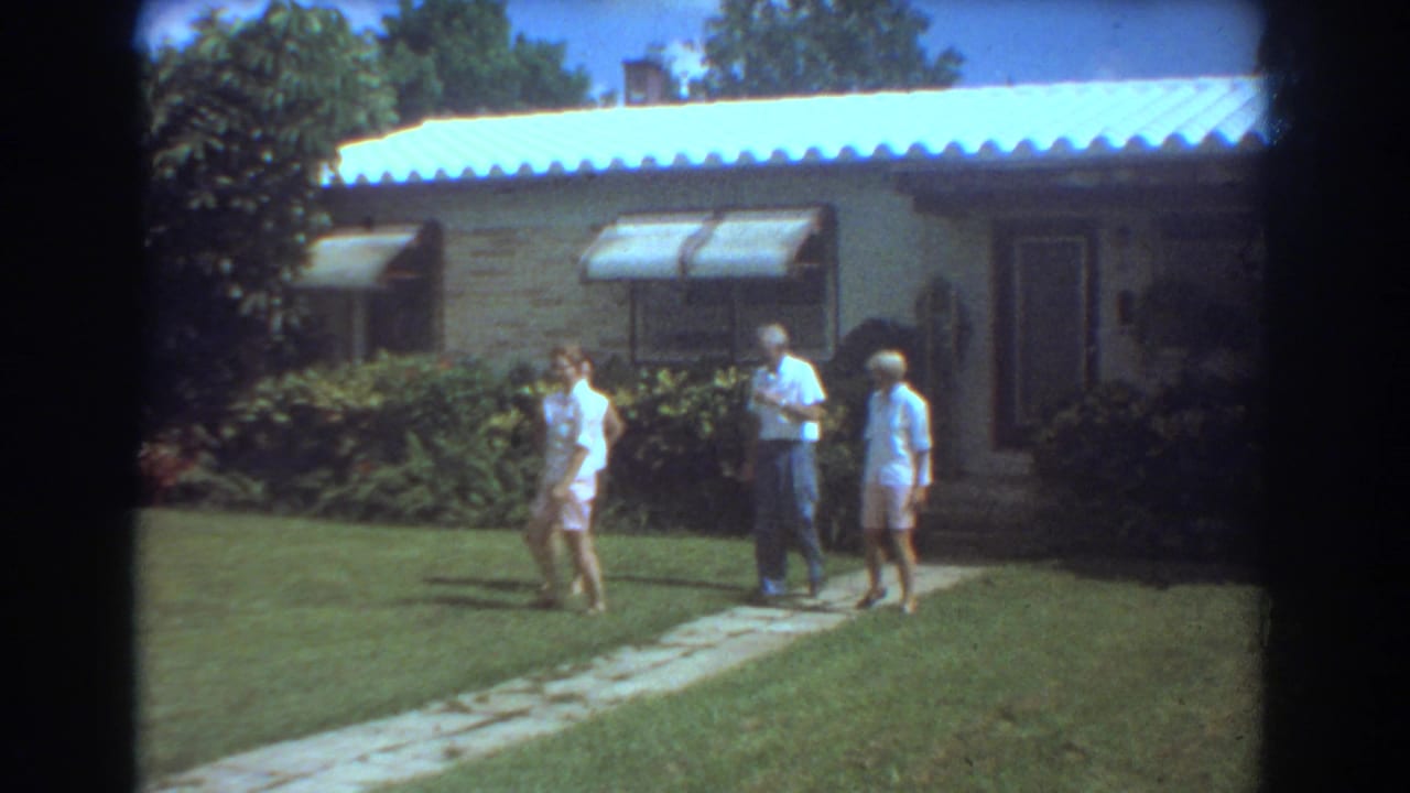 Three people continue walking in the Fort Lauderdale front yard, with a bright roofline, awnings, shrubs, and doorway visible behind them.