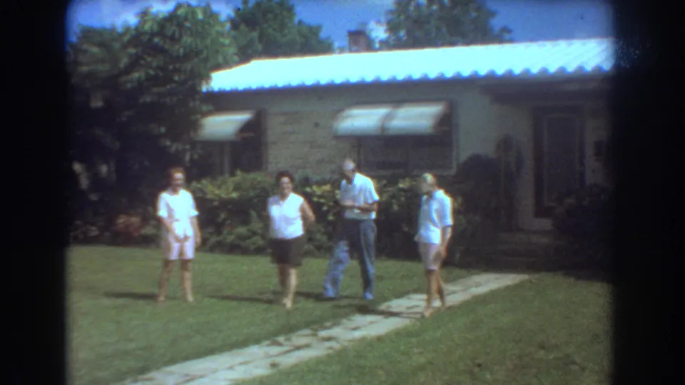 In 1967 Fort Lauderdale, four older adults move casually across a suburban yard outside a brick house with awnings.