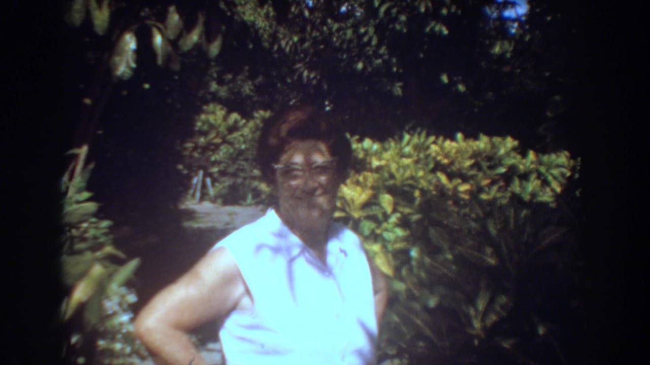In 1967 Fort Lauderdale home-movie footage, a woman in glasses and a white sleeveless shirt faces the camera in a lush garden.