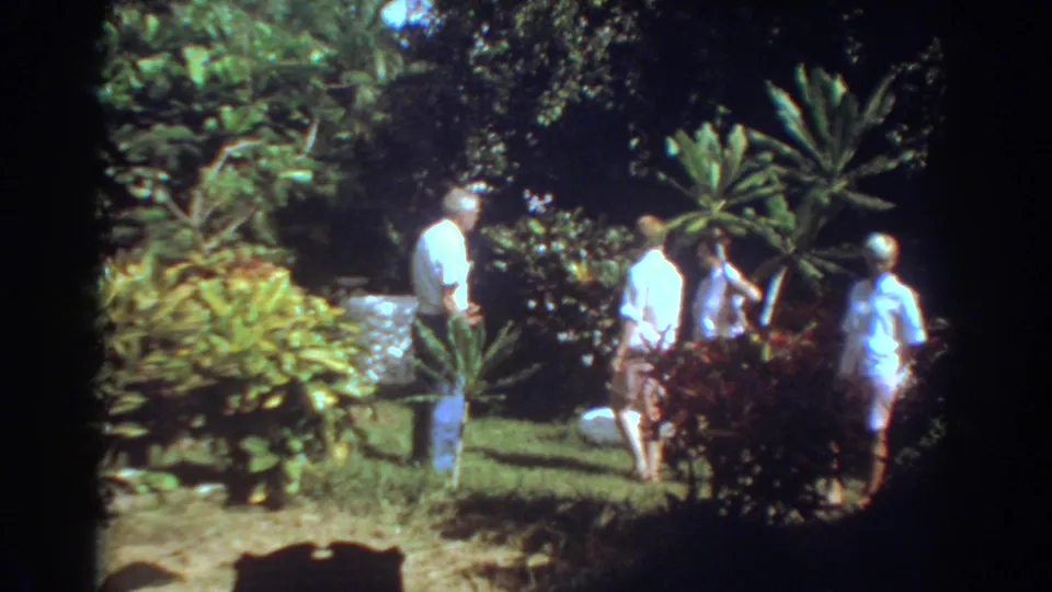 In 1967 Fort Lauderdale footage, adults in light clothing gather in a tropical garden with dense foliage and a lawn.