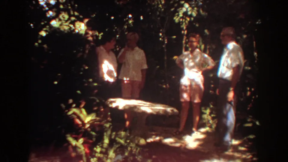A Fort Lauderdale 1967 home-movie frame shows adults gathered around a pale stone table under dense tree cover.