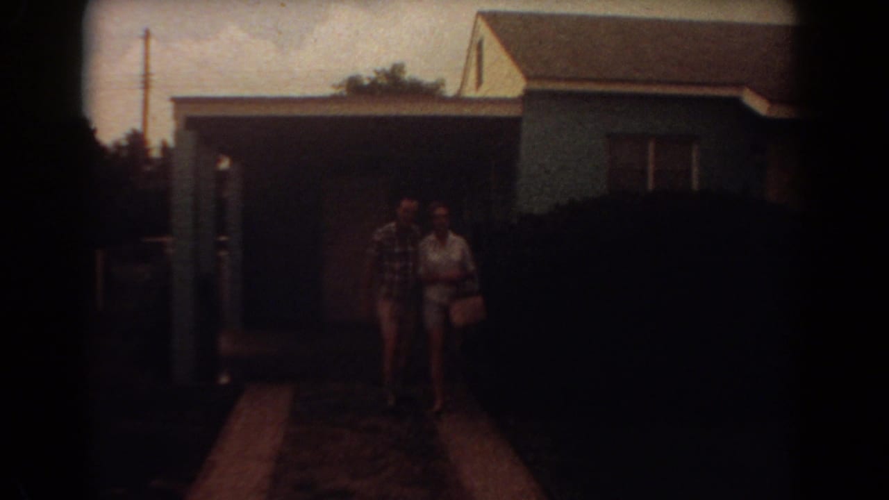 Adult couple stands on a driveway in front of a blue house and dark shrub.