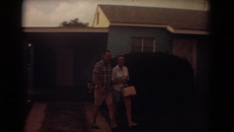 The couple approaches the camera on a driveway beside a large shrub and blue house in Fort Lauderdale, Florida.