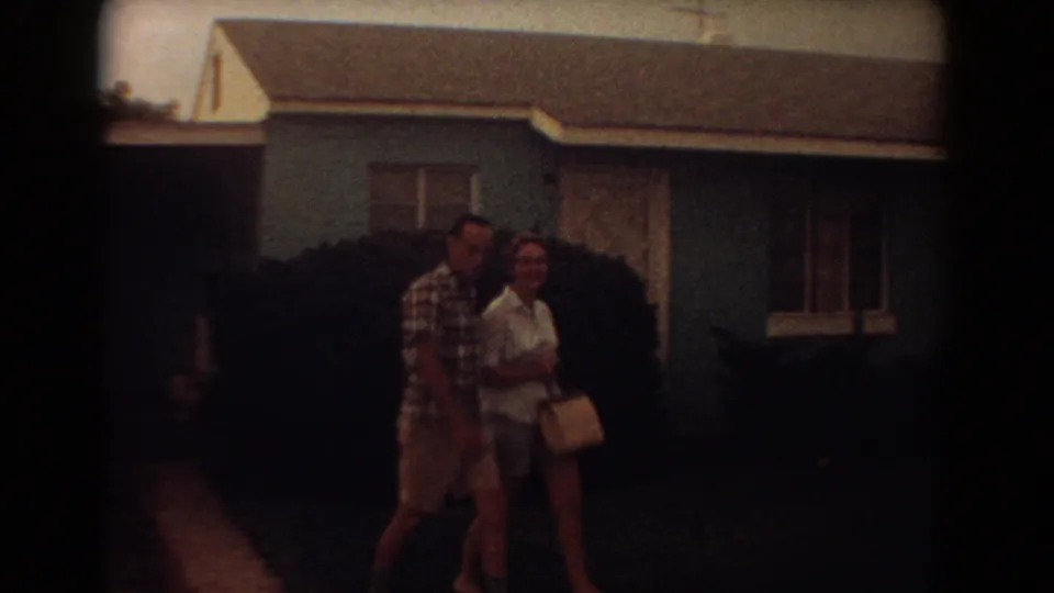 Near the end of the shot, the couple walks close to the camera, with the blue house and shrub behind them.