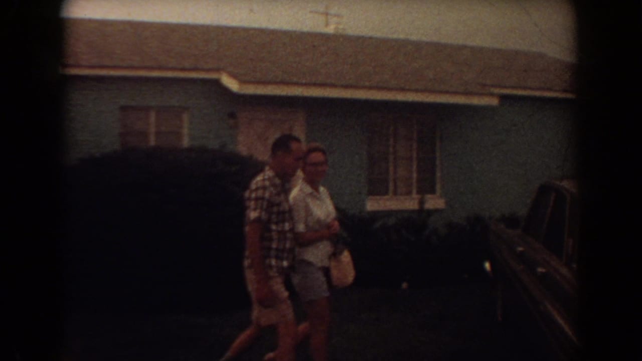Couple walking across a front yard beside parked cars and a light blue house.