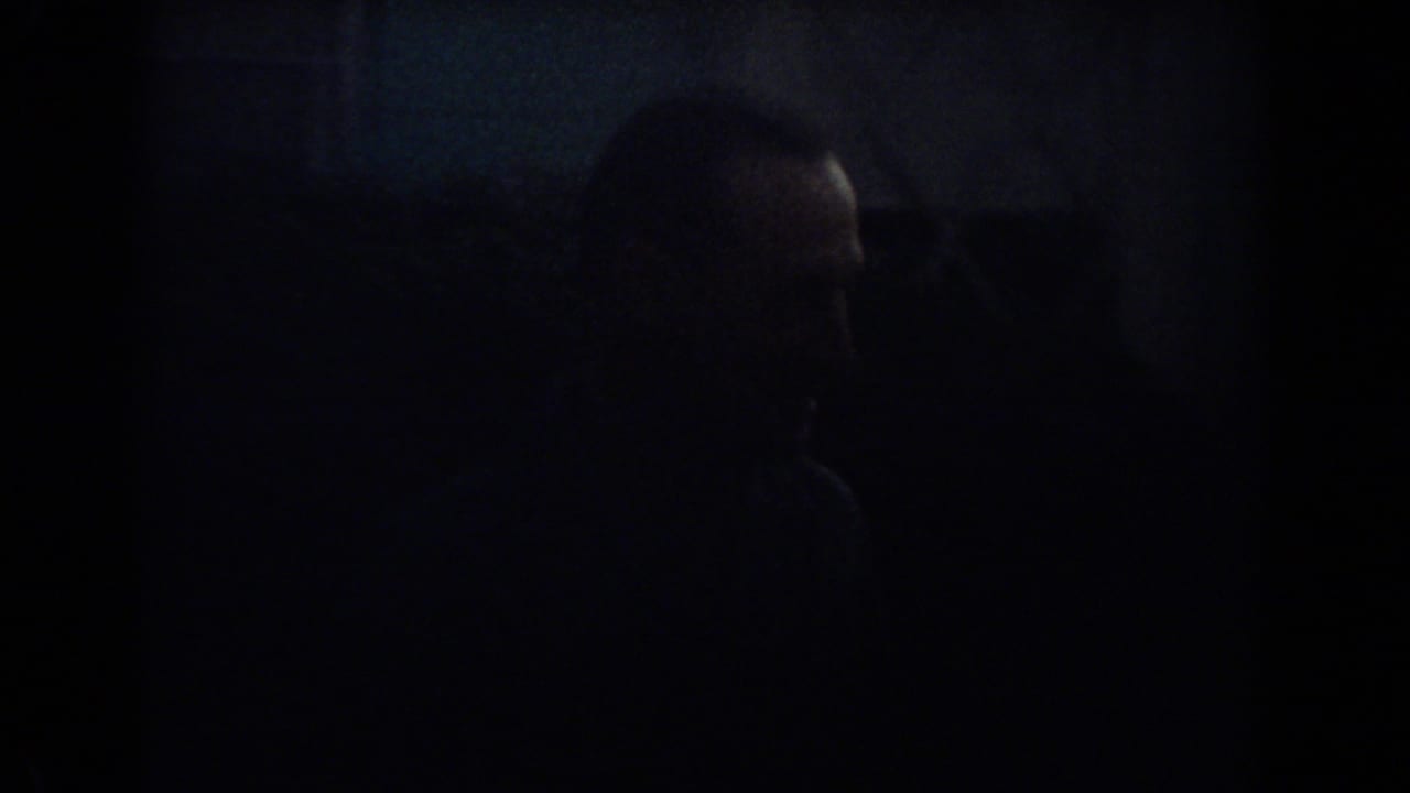 Dark frame with a man partly visible near a car or garage area.
