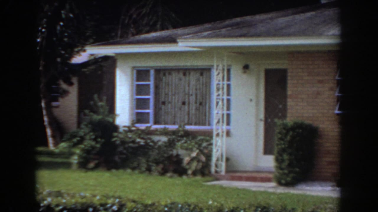 Cream and brick single-story house with front door, window, shrubs, lawn, and palm shadows.