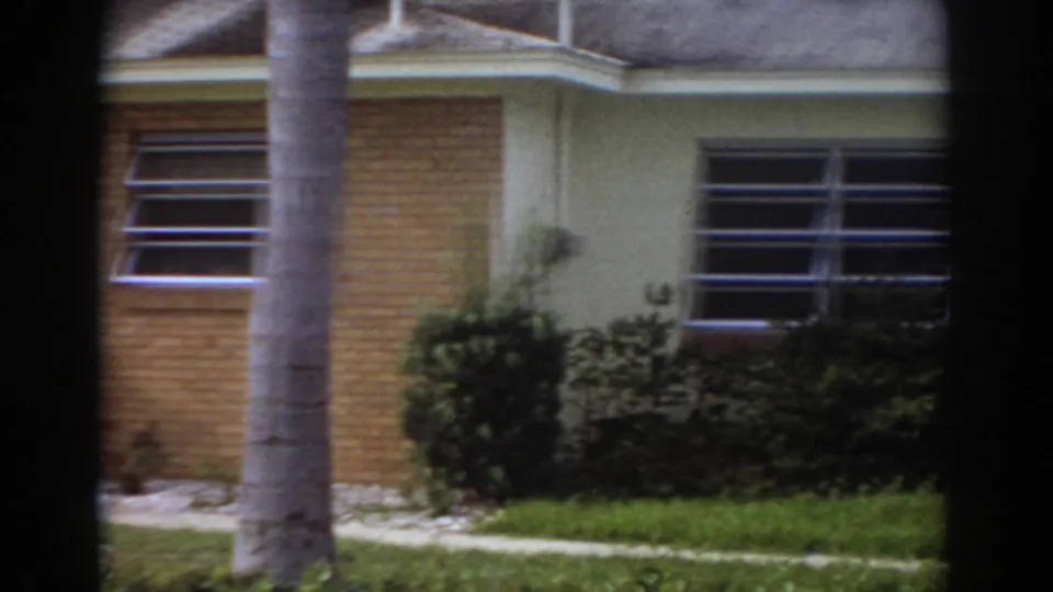 1967 Fort Lauderdale exterior frame showing louvered windows, a brick-and-cream wall, low landscaping, grass, walkway, and a palm trunk.