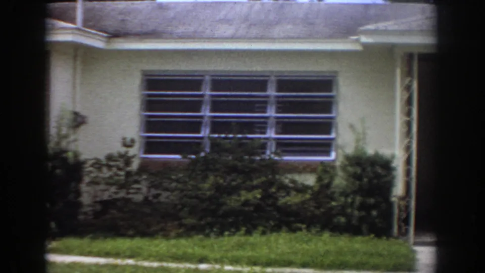 1967 Fort Lauderdale frame centered on a large louvered window, cream exterior wall, shrubs, grass, roofline, and a partial doorway edge.