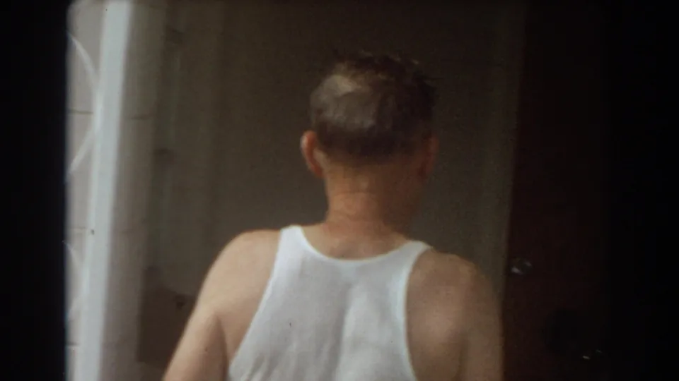 An older man in a white tank top turns away from the camera and steps into a dim residential interior.
