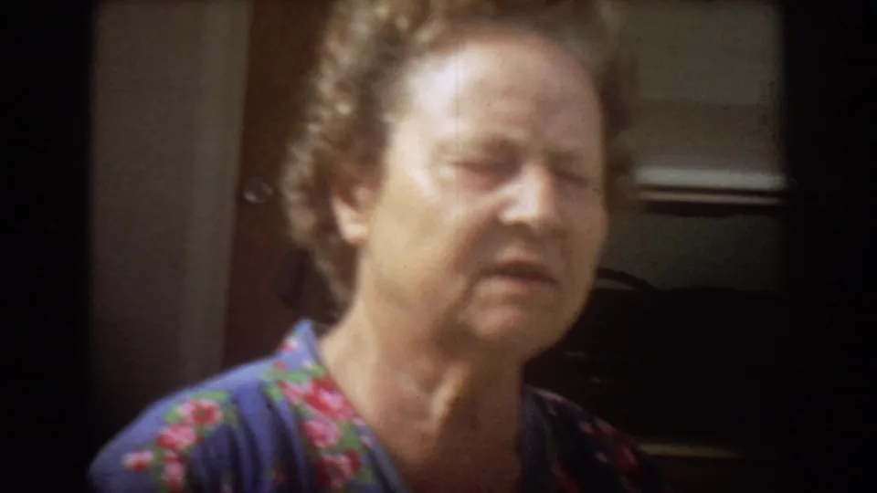 An older woman in a blue floral blouse appears in close view, filmed informally in a residential setting.