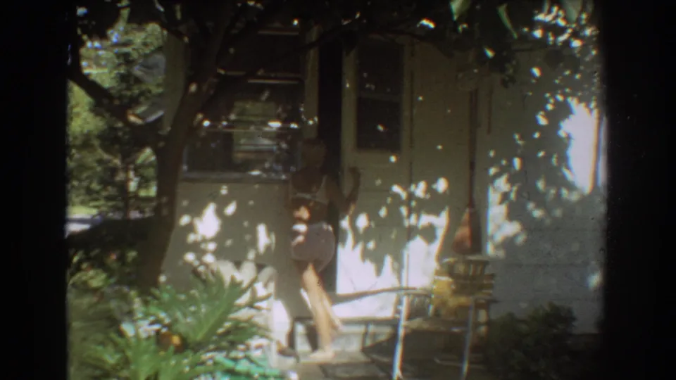 Young woman in a light swimsuit steps from the doorway beside porch plants at a white Fort Lauderdale bungalow.