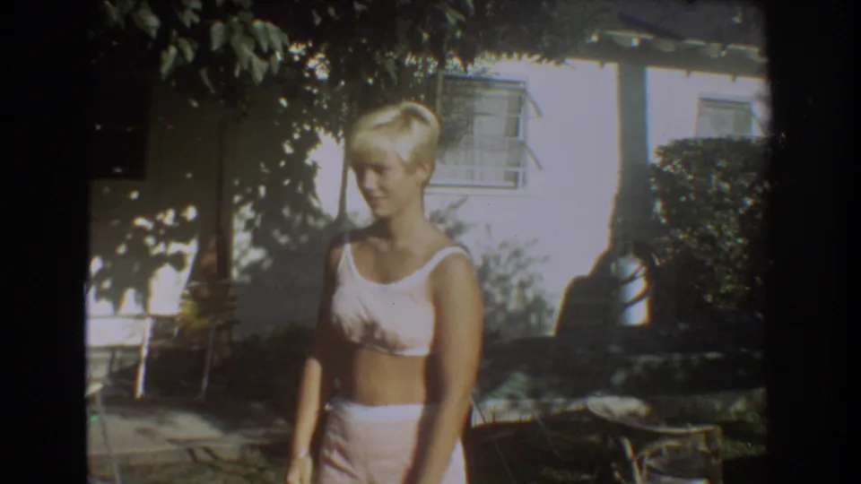 In Fort Lauderdale home-movie footage from 1967, a blonde woman in a white swimsuit stands near a house and patio furniture.
