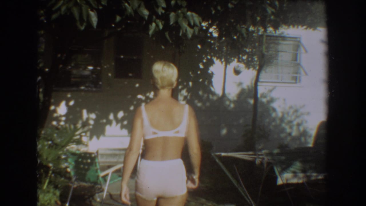 The woman in a white two-piece swimsuit walks away from camera through a Fort Lauderdale backyard in 1967 footage.