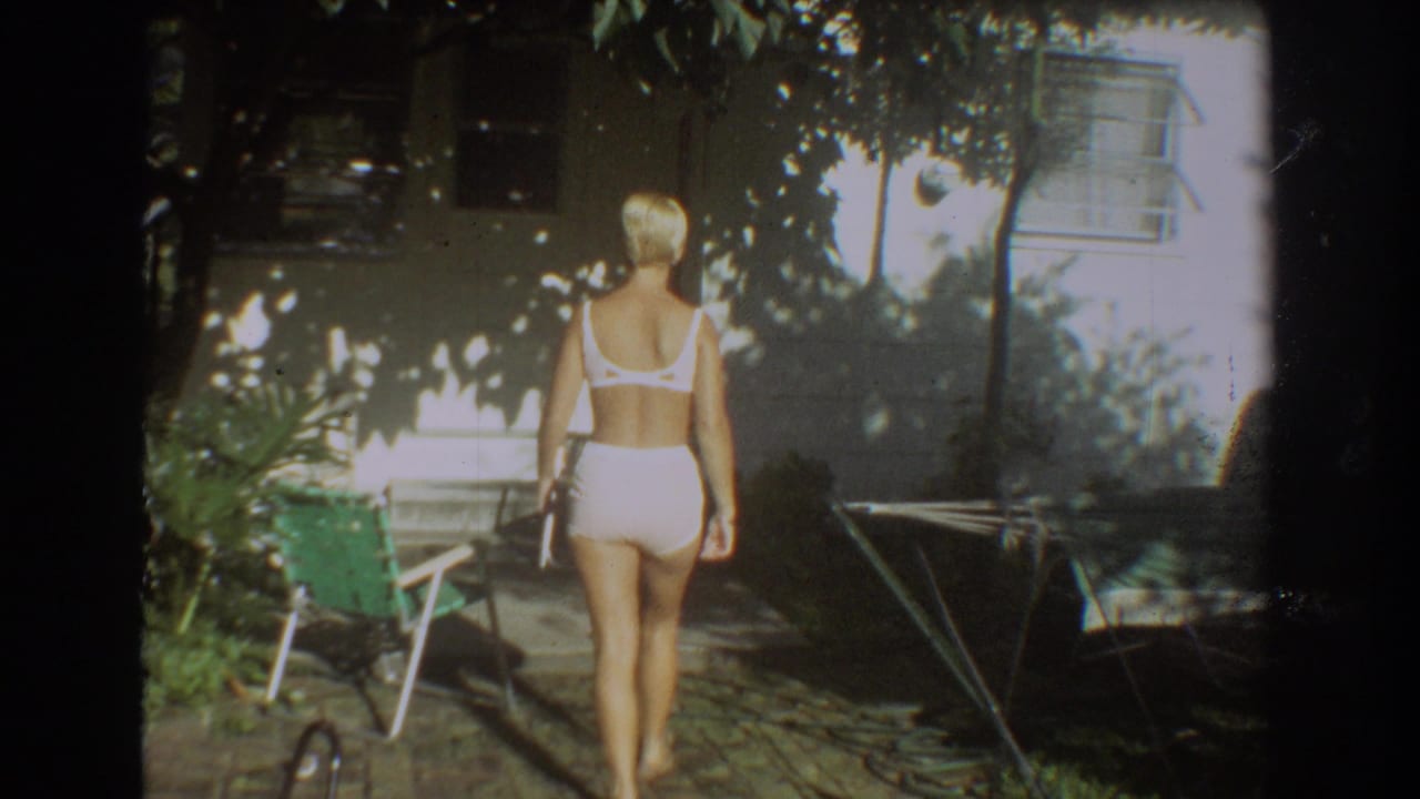 A rear view shows the woman crossing the 1967 Fort Lauderdale backyard past a green chair, plants, and a light house exterior.