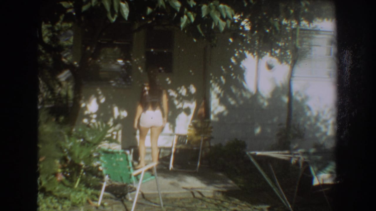 Woman in a white swimsuit walks near lawn chairs beside a shaded house.