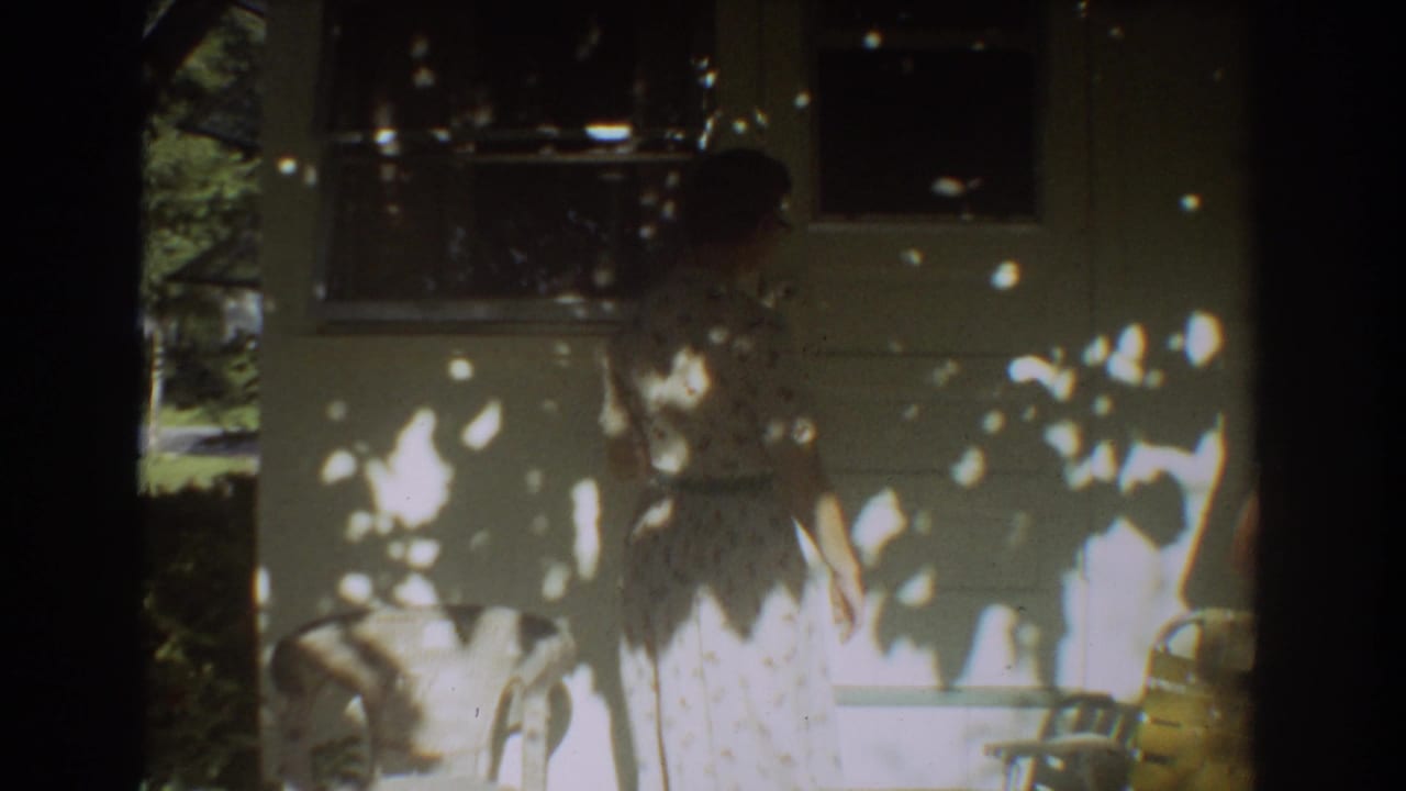 Woman in a floral dress stands on a porch near windows and chairs.