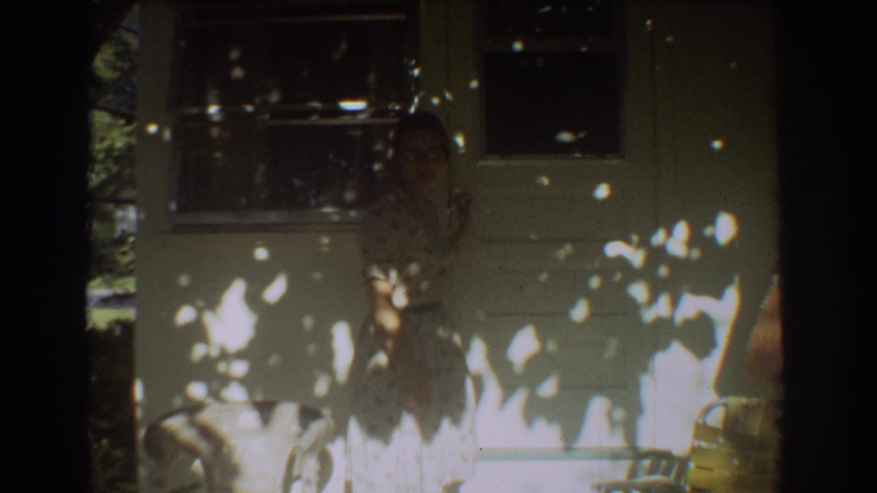 The frame shows a woman in a floral dress near the house exterior, with tree shadows and soft 8mm home-movie focus.